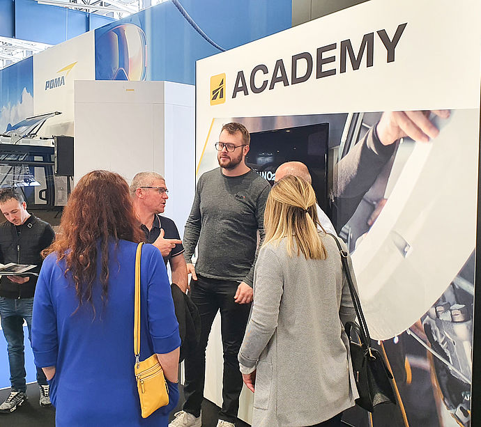 Trade show visitors conversing at TechnoAlpin Academy stand with screen and training graphics.