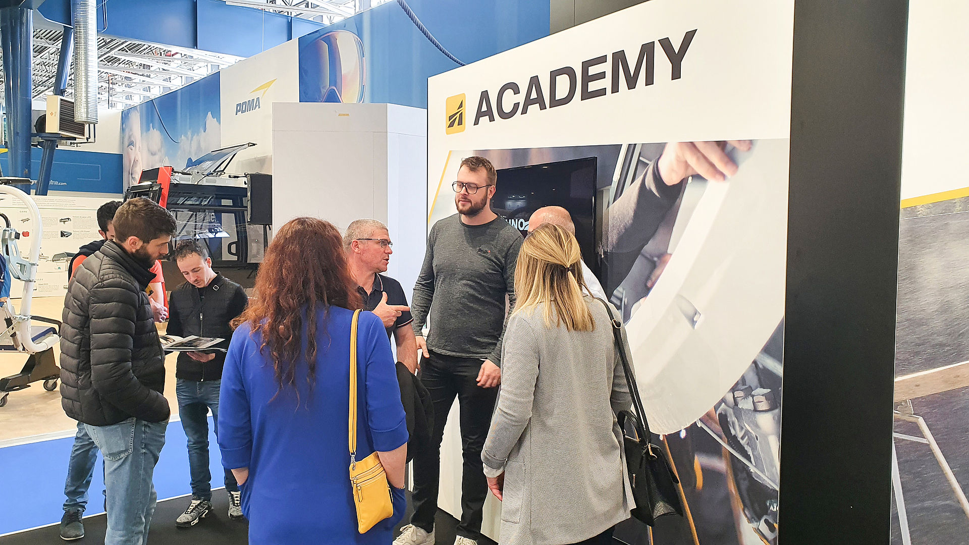 Trade show visitors conversing at TechnoAlpin Academy stand with screen and training graphics.