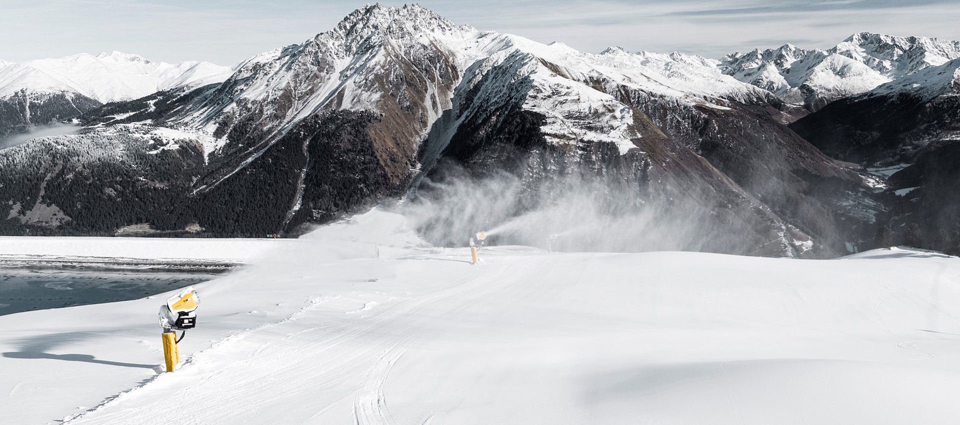 Schneeerzeuger beschneit Piste mit Blick auf Bergsee und Alpenpanorama