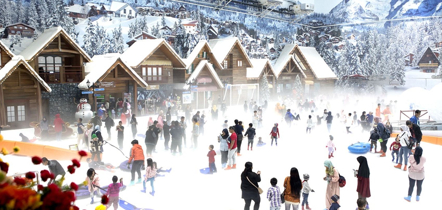 Visitors in indoor snow park with wooden huts and snowy mountain scenery