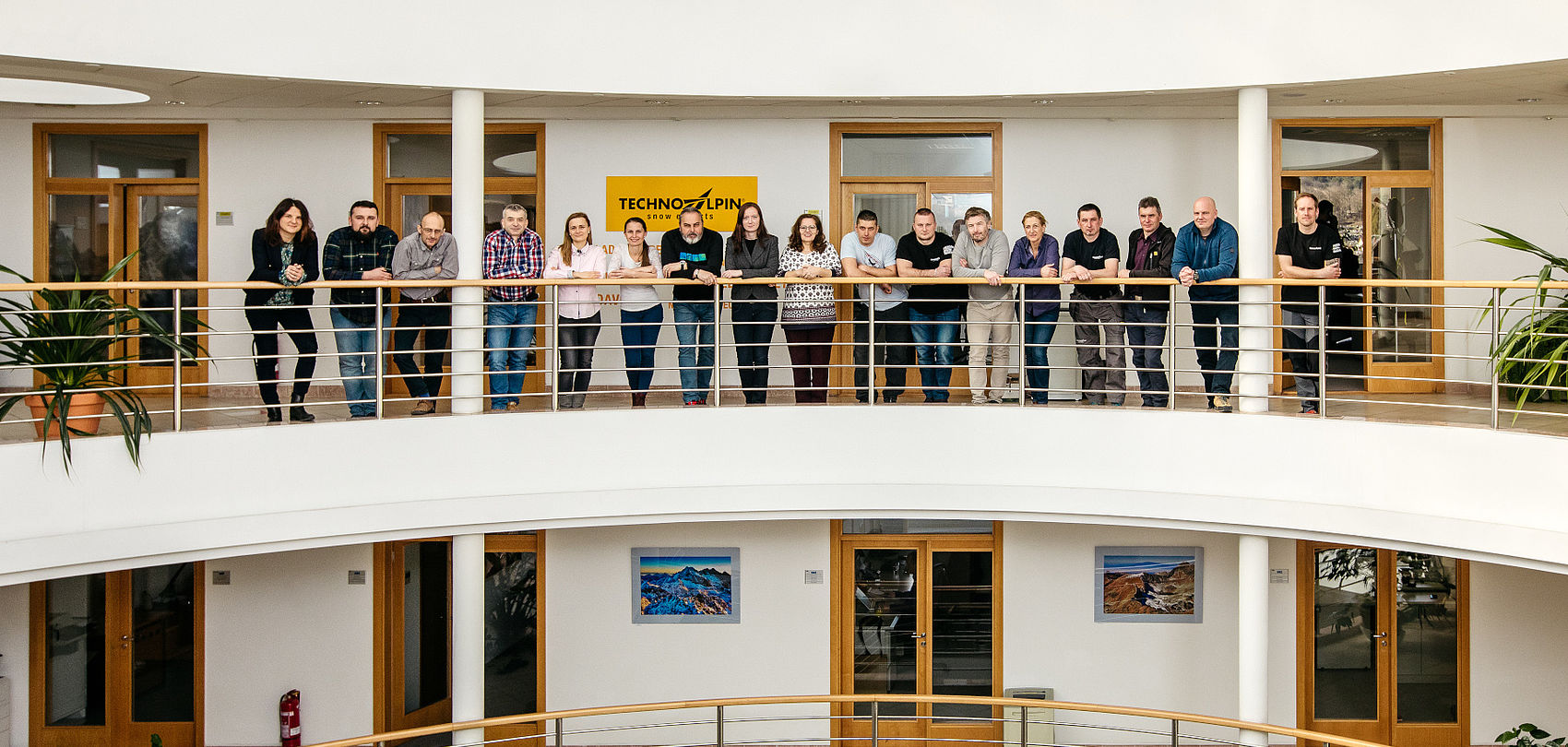 TechnoAlpin Mitarbeitergruppe posiert an der geschwungenen Balkonbrüstung in einem modernen Bürogebäude vor dem Firmenlogo