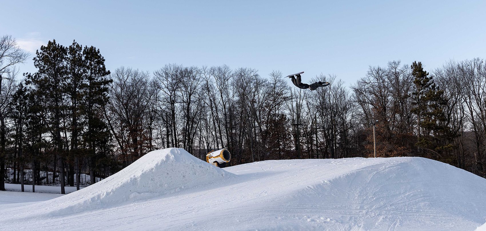 Freestyle snowboarder performing a jump, with a TechnoAlpin TF10 in the background