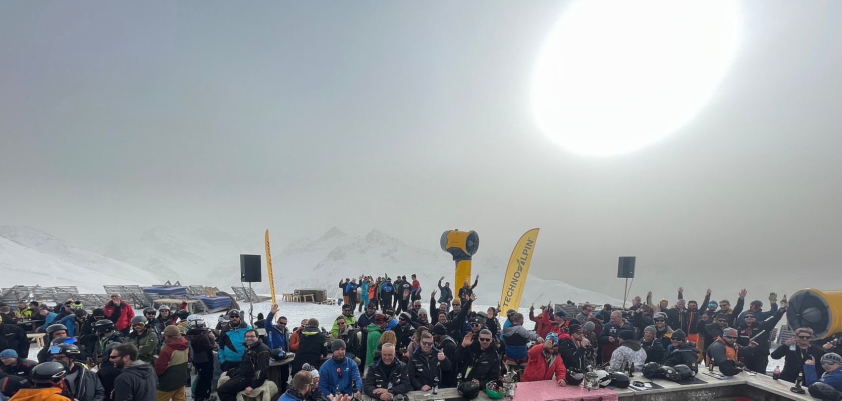 Grande folla all'evento TechnoAlpin su terrazza montana con bandiere gialle del marchio.