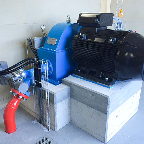 Blaue Pelton-Turbine mit schwarzem Elektromotor auf Betonsockel in einer Wasserkraftwerksanlage.