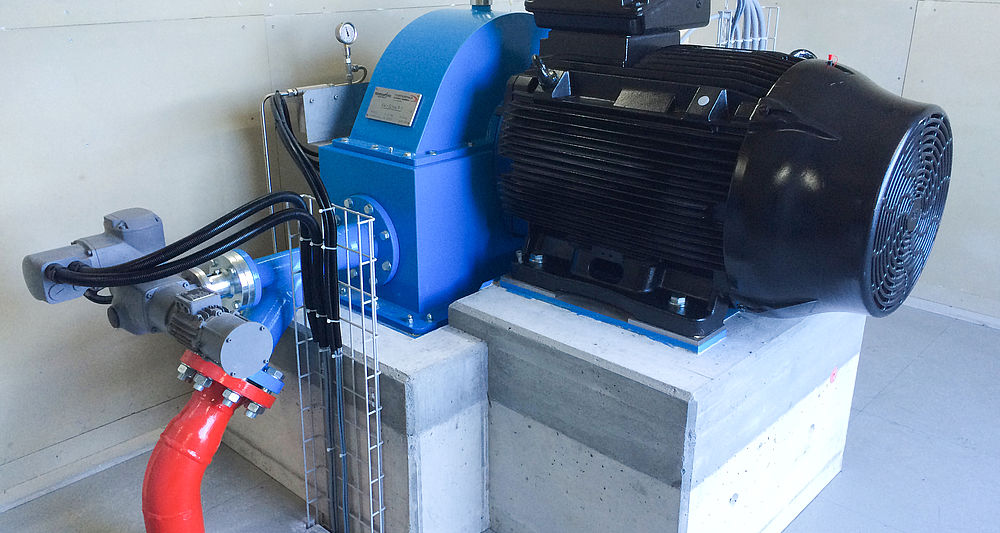 Turbina Pelton blu con motore elettrico nero su piedistallo in cemento in impianto idroelettrico.