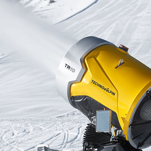 Nahaufnahme eines gelben TR10 Schneeerzeugers mit Ventilator vor verschneitem Pistenhintergrund.