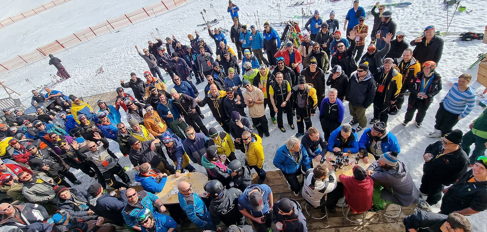 Große Gruppe von Personen in Skikleidung versammelt auf einer Terrasse im Schnee, viele winken in die Kamera