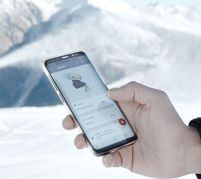 Hand hält Smartphone mit Steuerungs-App vor TechnoAlpin Schneeerzeuger im Bergpanorama