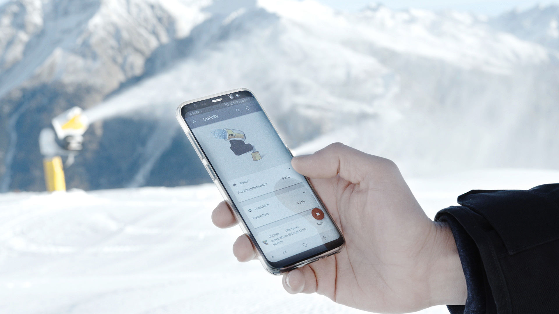 Hand hält Smartphone mit Steuerungs-App vor TechnoAlpin Schneeerzeuger im Bergpanorama