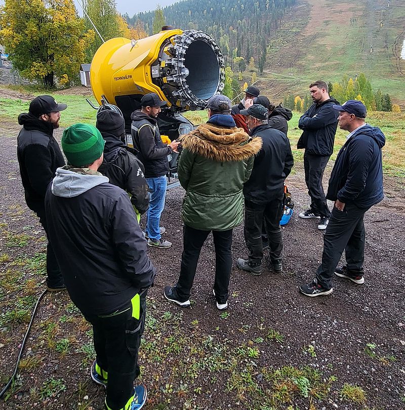 Gruppe bei Academy on Tour Schulung an gelbem Schneeerzeuger vor herbstlicher Berglandschaft
