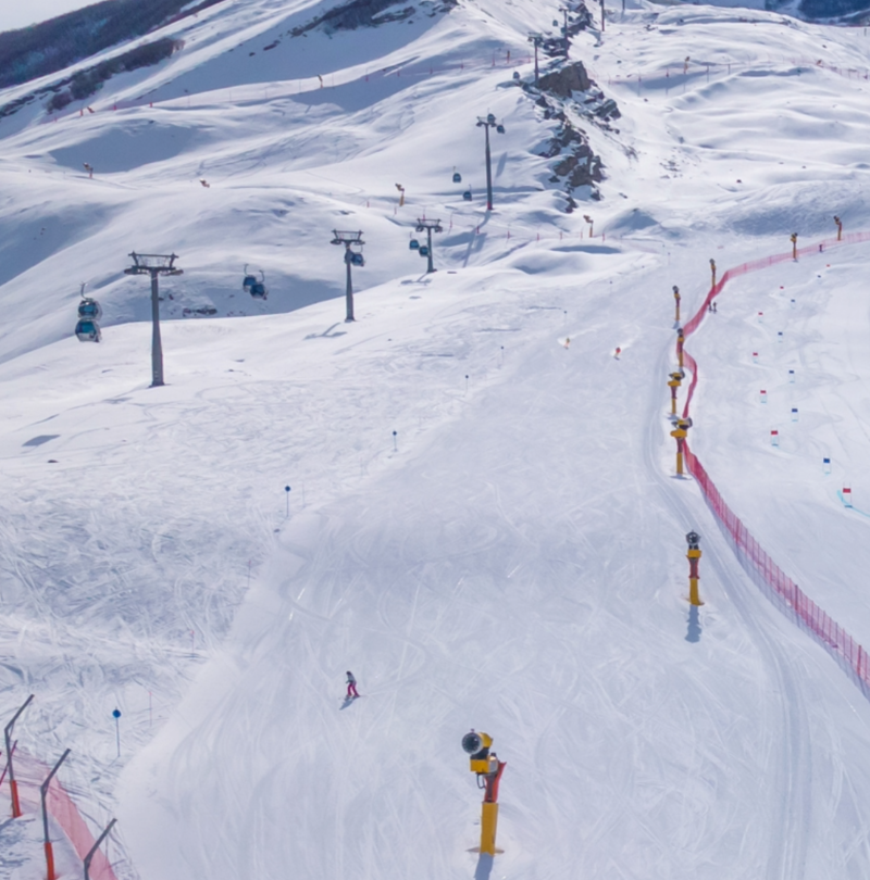 Vue aérienne piste damée avec enneigeurs TechnoAlpin jaunes, télésiège et crête montagneuse