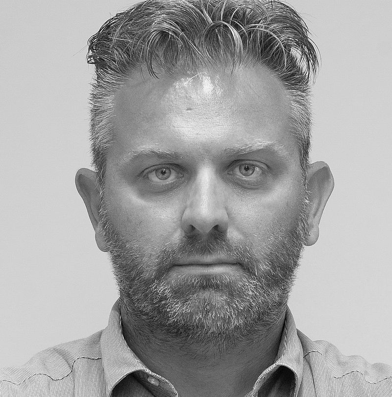 Omar Faustini, TechnoAlpin Mitarbeiter, Schwarz-Weiß-Portrait mit grau-dunklem Haar, kurzem Bartansatz und neutralem Ausdruck in kariertem Hemd