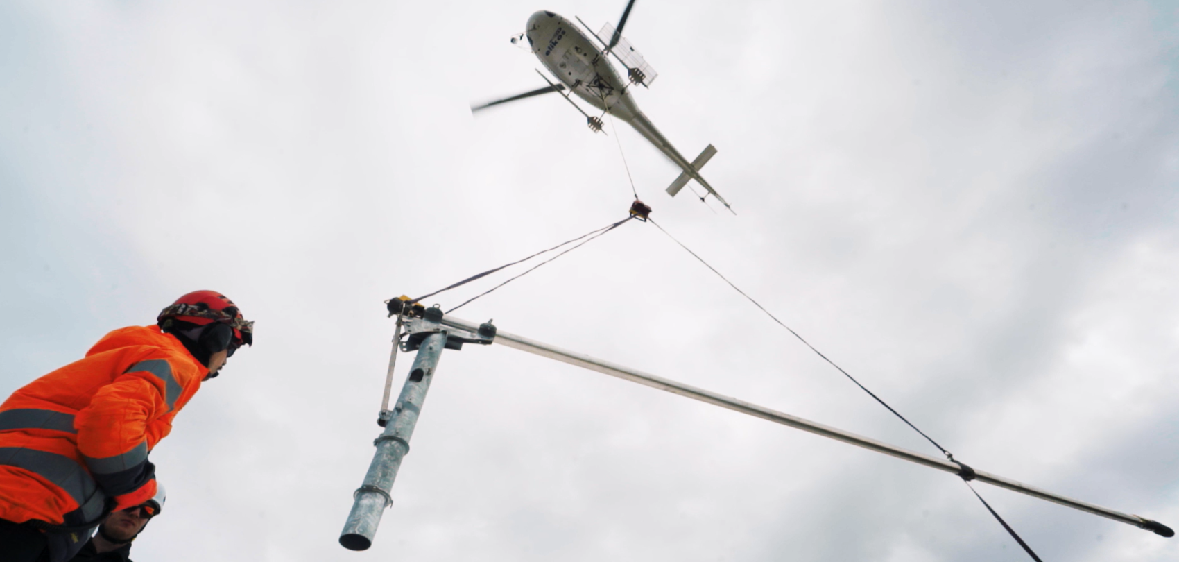 Helicopter transports snow lance, construction worker watches installation from below