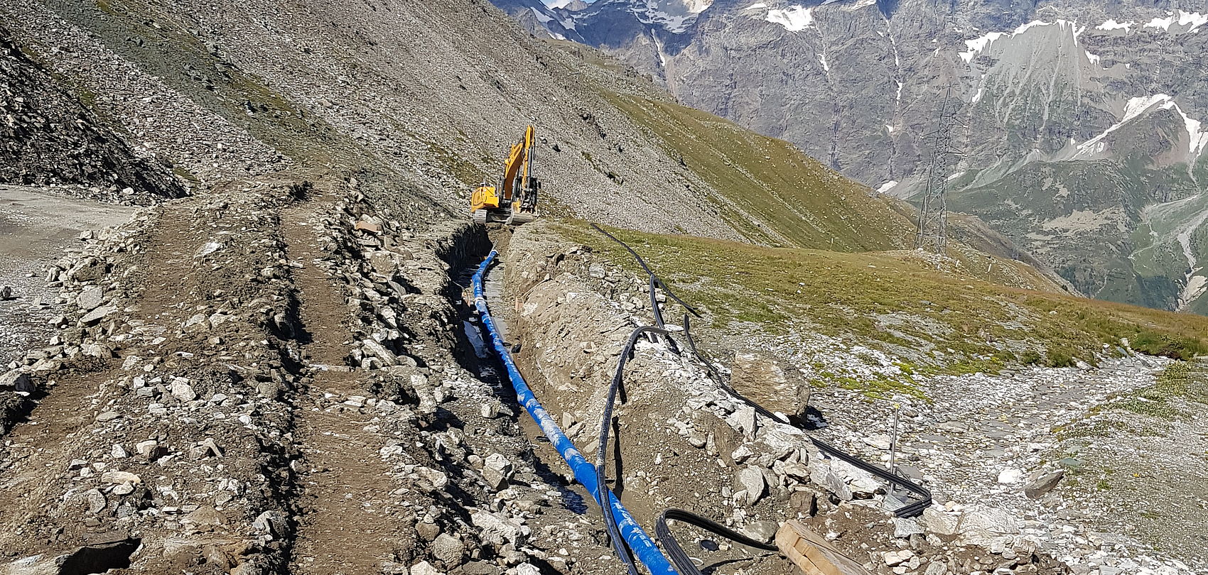 Verlegung blauer Wasserleitungen im Hochgebirge mit Bagger, Felshang und Bergpanorama im Hintergrund