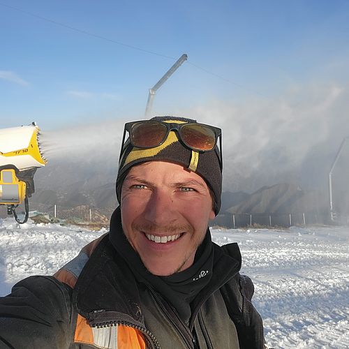 Lächelnder Mann mit Mütze und Sonnenbrille macht Selfie im Schnee neben gelbem TF10-Schneeerzeuger.