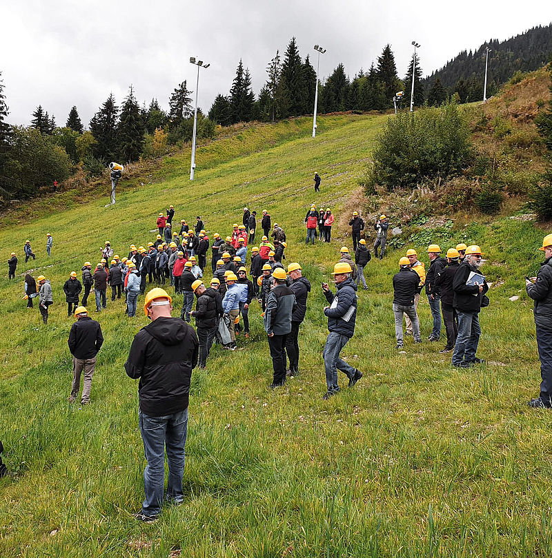 Large group wearing yellow helmets at TechnoAlpin on-mountain demonstration on grassy ski slope