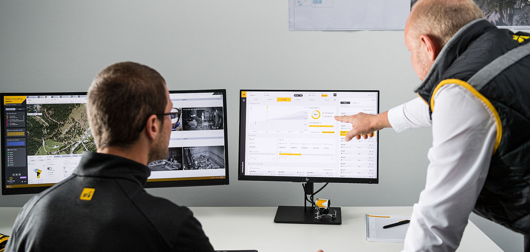 Two TechnoAlpin colleagues discussing Snowmaster analytics at monitor with miniature yellow snow gun model on desk