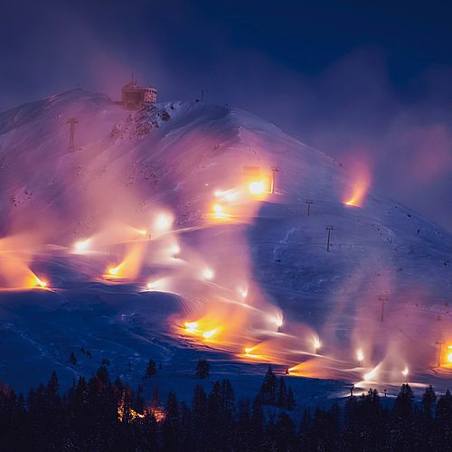 Beschneiungsanlagen im Nachtbetrieb an einem Berghang mit orange leuchtenden Schneeerzeugern.