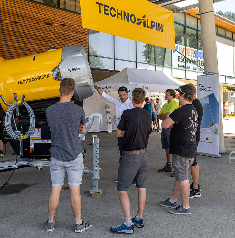 Visitor group inspecting yellow TechnoAlpin TR9 propeller snow gun at roadshow.