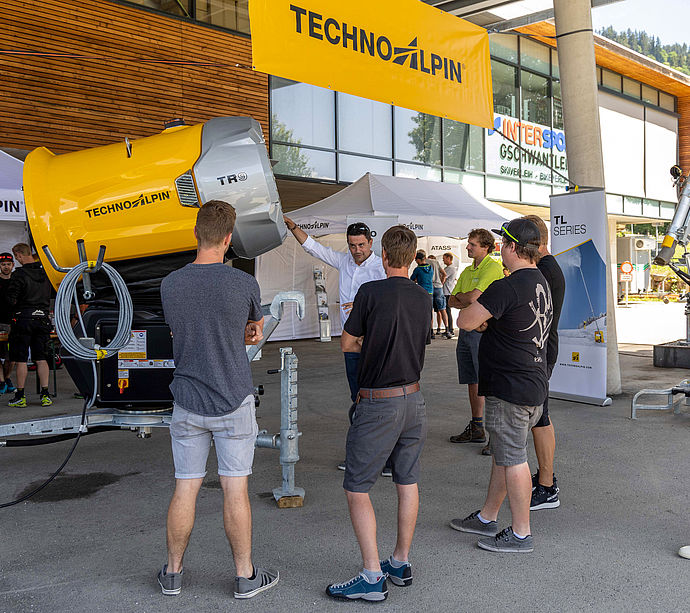 Visitor group inspecting yellow TechnoAlpin TR9 propeller snow gun at roadshow.