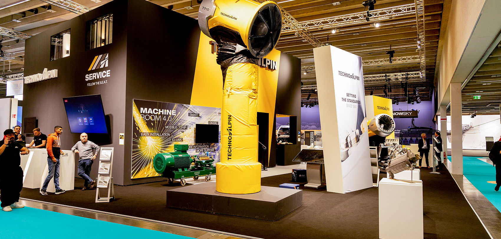 TechnoAlpin Interalpin 2023 Messestand mit gelber TT-Schneekanone, Machine Room 4.0 Bildschirm und Besuchern auf türkisem Teppich