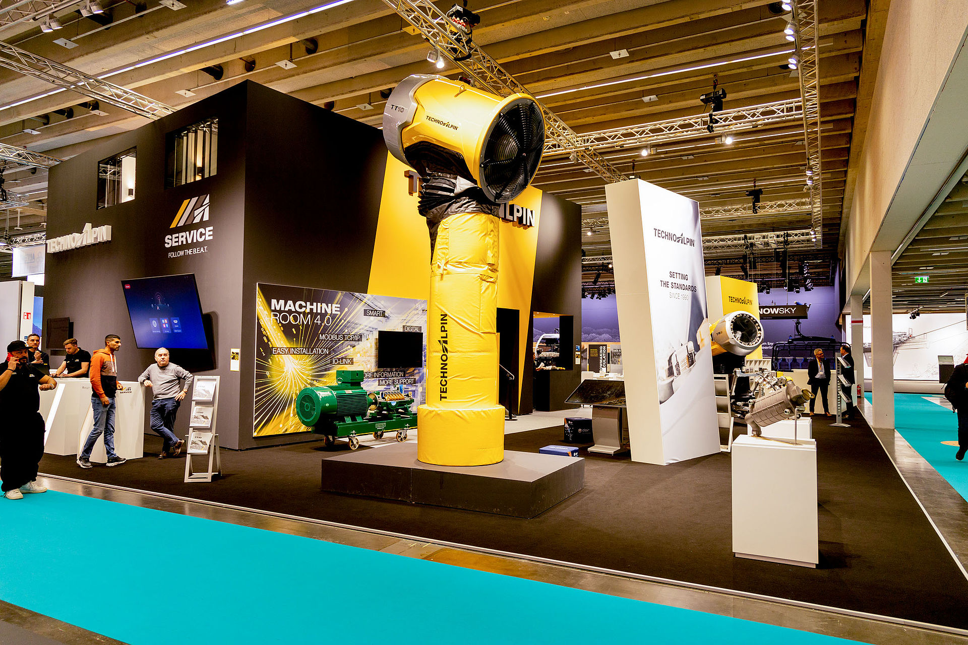 TechnoAlpin Interalpin 2023 Messestand mit gelber TT-Schneekanone, Machine Room 4.0 Bildschirm und Besuchern auf türkisem Teppich