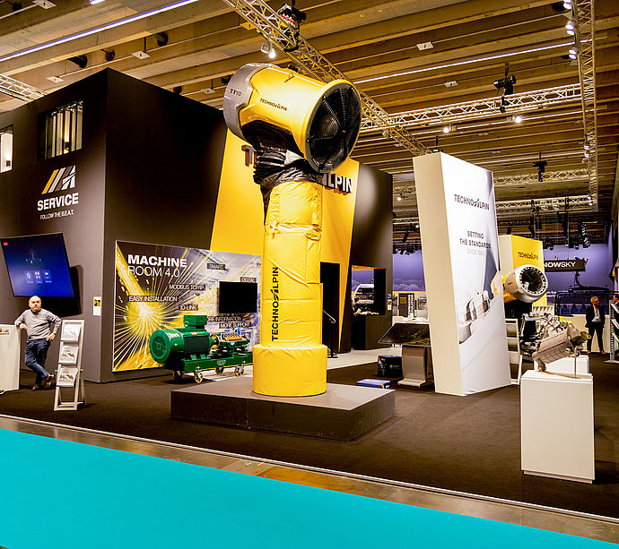 TechnoAlpin Interalpin 2023 Messestand mit gelber TT-Schneekanone, Machine Room 4.0 Bildschirm und Besuchern auf türkisem Teppich