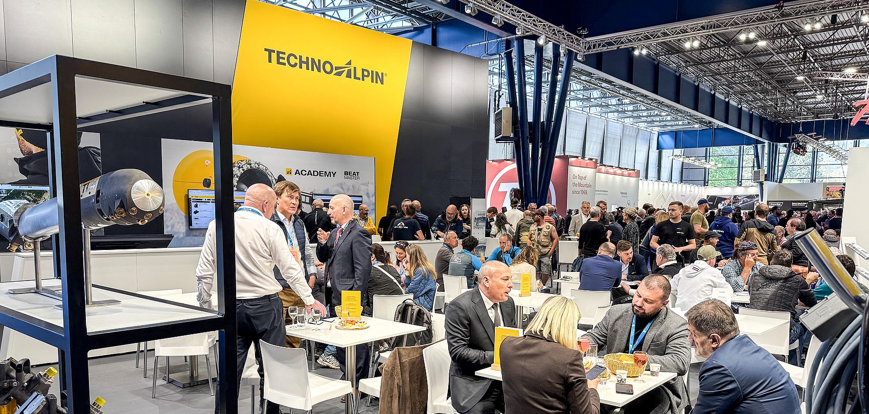 TechnoAlpin-Messestand mit gelbem Logo-Banner, Gäste sitzen an Tischen im Gespräch
