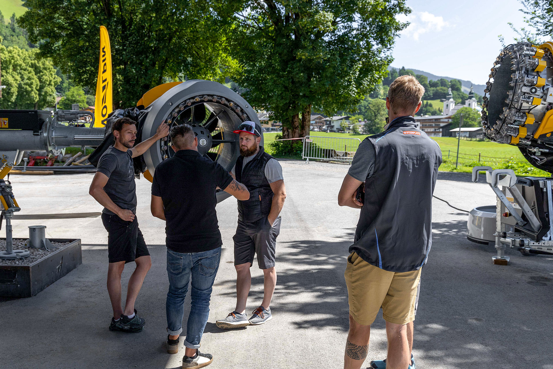 Men conversing between two propeller snow generators in shade with church tower in background.