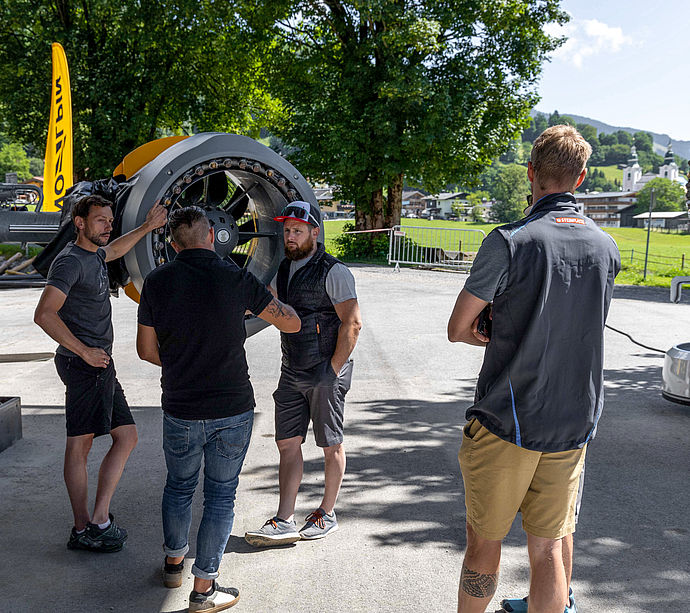 Men conversing between two propeller snow generators in shade with church tower in background.