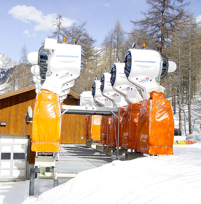 Row of white fan guns with orange shells in front of wooden building at ski resort