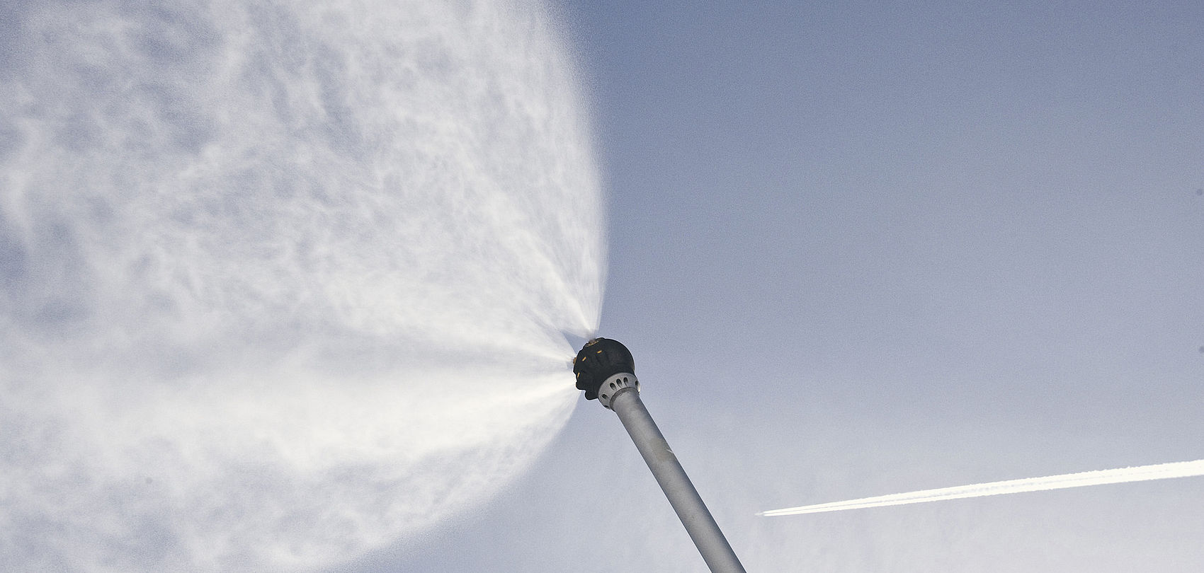 TechnoAlpin Schneelanzenkopf von unten, sprüht feinen Wassernebel in breitem Fächer gegen blauen Himmel mit Kondensstreifen