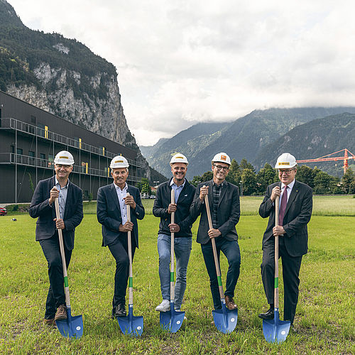 Fünf Männer mit Bauhelmen und Schaufeln auf grüner Wiese vor Tellpark-Gebäude und Bergkulisse.