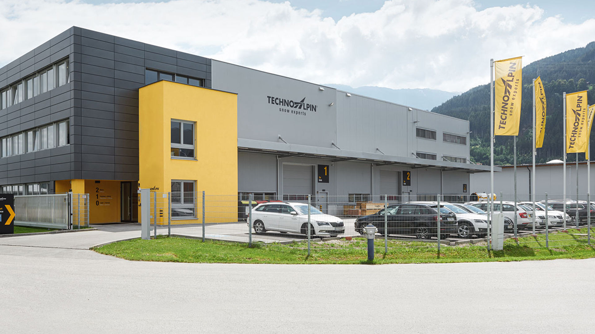 TechnoAlpin Firmengebäude mit gelben Markenfahnen und Logo an grauer Fassade in Südtirol
