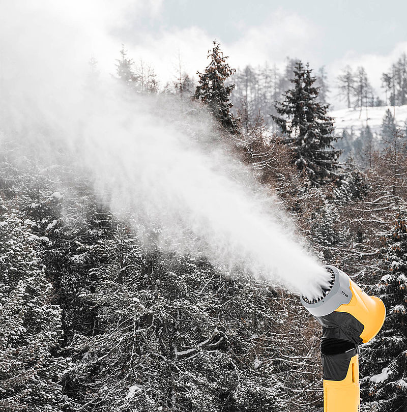 Gelbe TechnoAlpin Propellerkanone bei der Schneeproduktion vor verschneitem Nadelwald