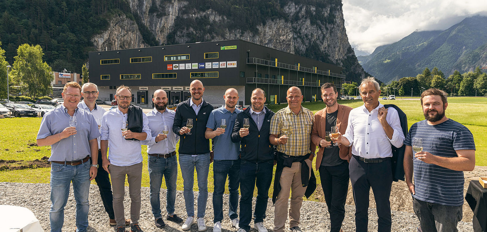 Group photo with champagne glasses at groundbreaking in front of Tellpark building and mountains.
