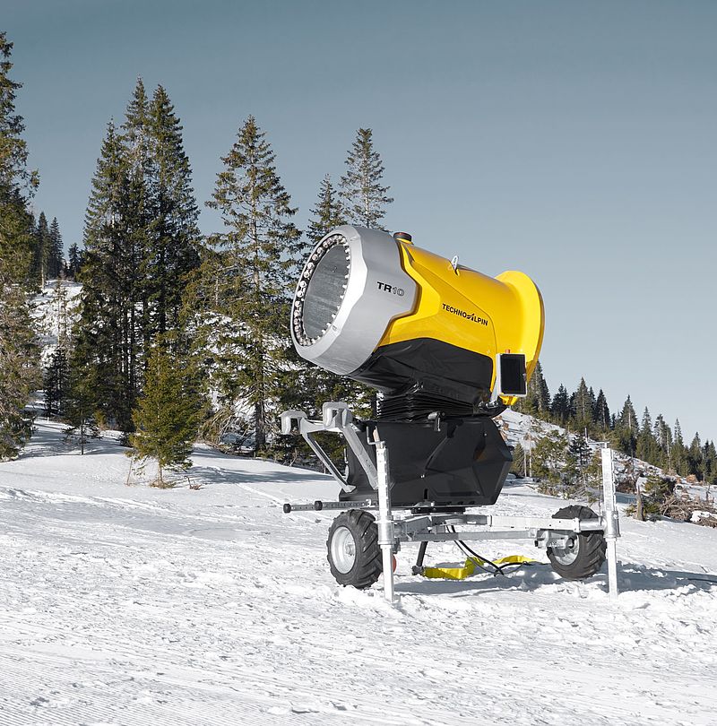 Gelbe TechnoAlpin TR10 Propellerschneeerzeuger auf präparierter Skipiste mit Nadelbäumen
