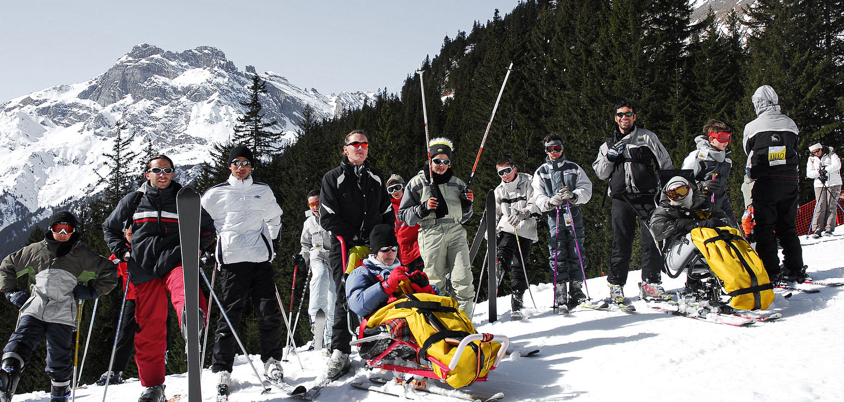 Group of skiers with disabled people in monoskis on snowy slope in front of mountain panorama.