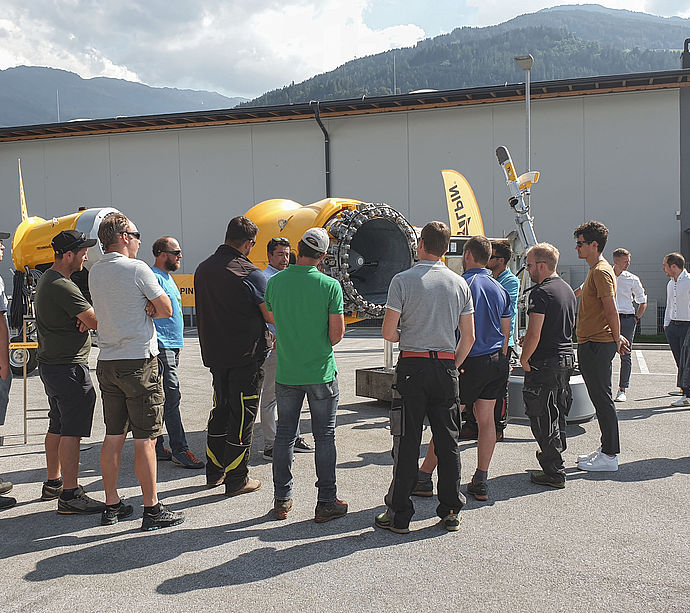 Gruppo visitatori davanti a cannone da neve TechnoAlpin TT10 all'esterno dello stabilimento di Volders con montagne
