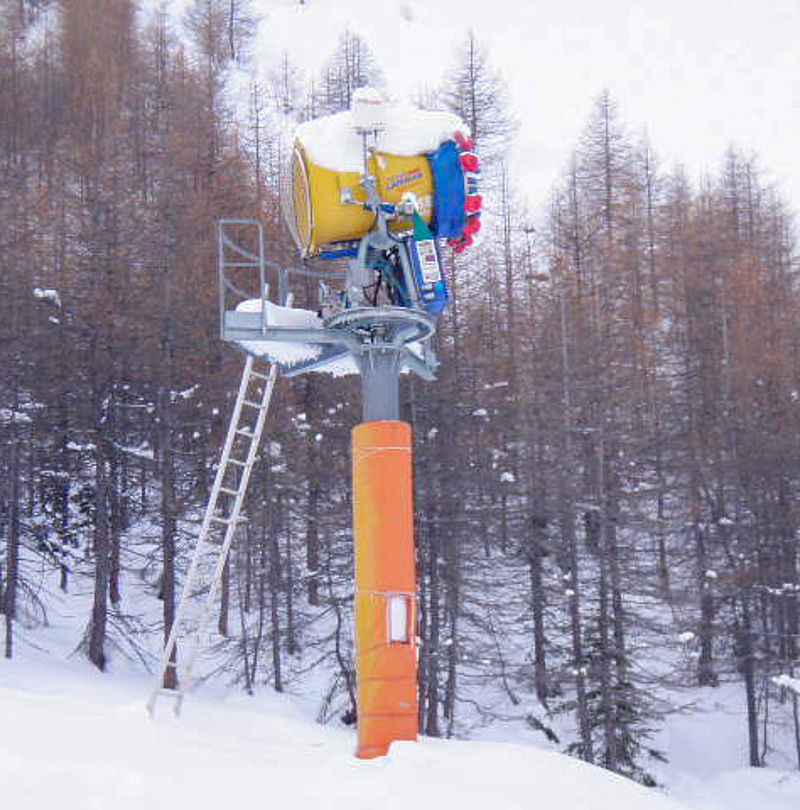 Snow gun with orange guard on mast in front of snowy larch forest
