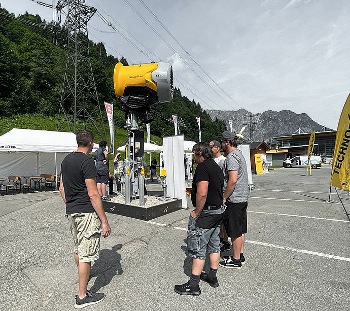 Visitors examining yellow TT10 snow gun on swivel arm at TechnoAlpin roadshow.