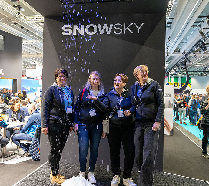 Vier Frauen lachen und freuen sich über künstlichen Schneefall der TechnoAlpin SNOWSKY Demonstrationseinheit beim Interalpin Messestand