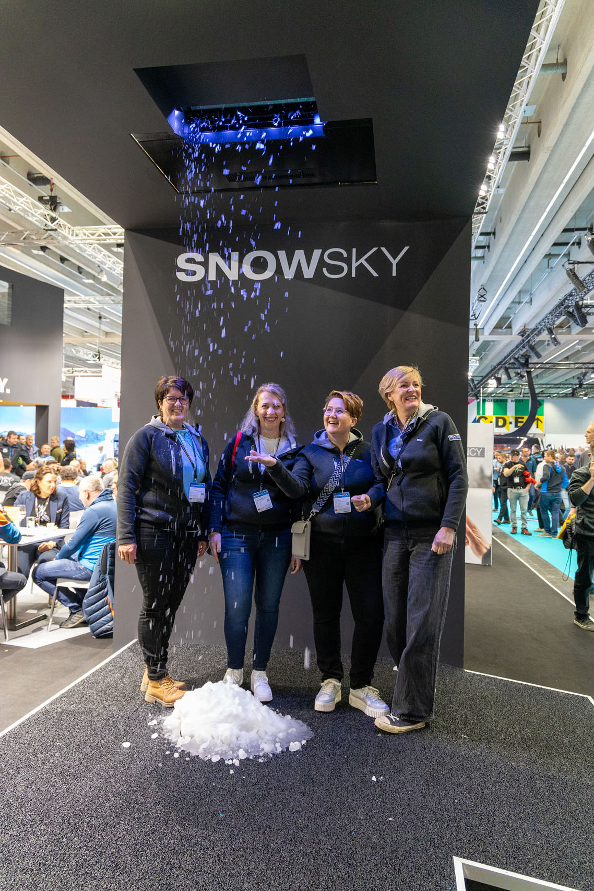 Vier Frauen lachen und freuen sich über künstlichen Schneefall der TechnoAlpin SNOWSKY Demonstrationseinheit beim Interalpin Messestand