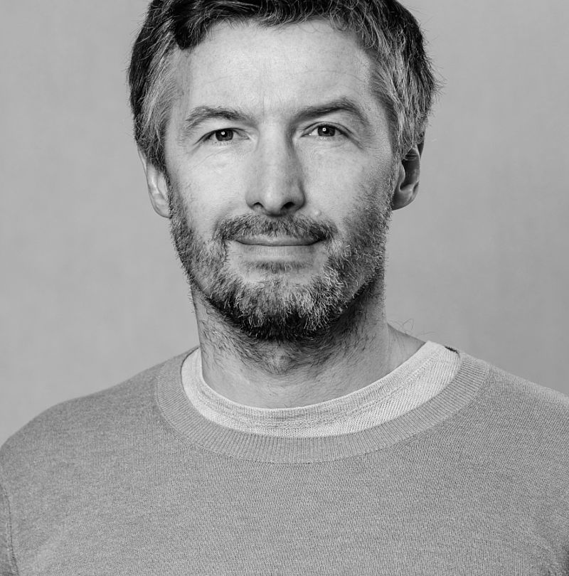 Roman Hotera, TechnoAlpin Mitarbeiter, Schwarz-Weiß-Portrait mit kurzem lockigem Haar, Bart und sanftem Lächeln in hellem Pullover