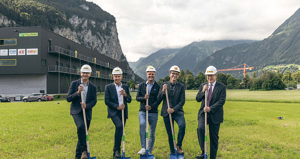 Fünf Personen mit Bauhelmen und Spaten beim Spatenstich auf grüner Wiese vor Bergkulisse.