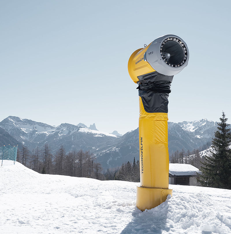 Yellow TechnoAlpin TT9 snow generator on ski slope in front of alpine panorama under blue sky.