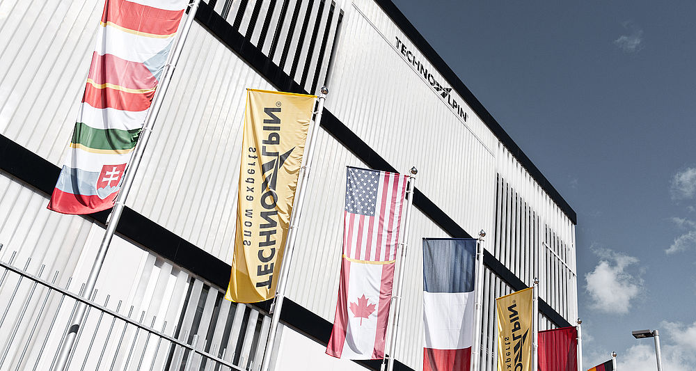 TechnoAlpin Firmengebäude mit internationalen Flaggen und gelben Markenfahnen vor blauem Himmel