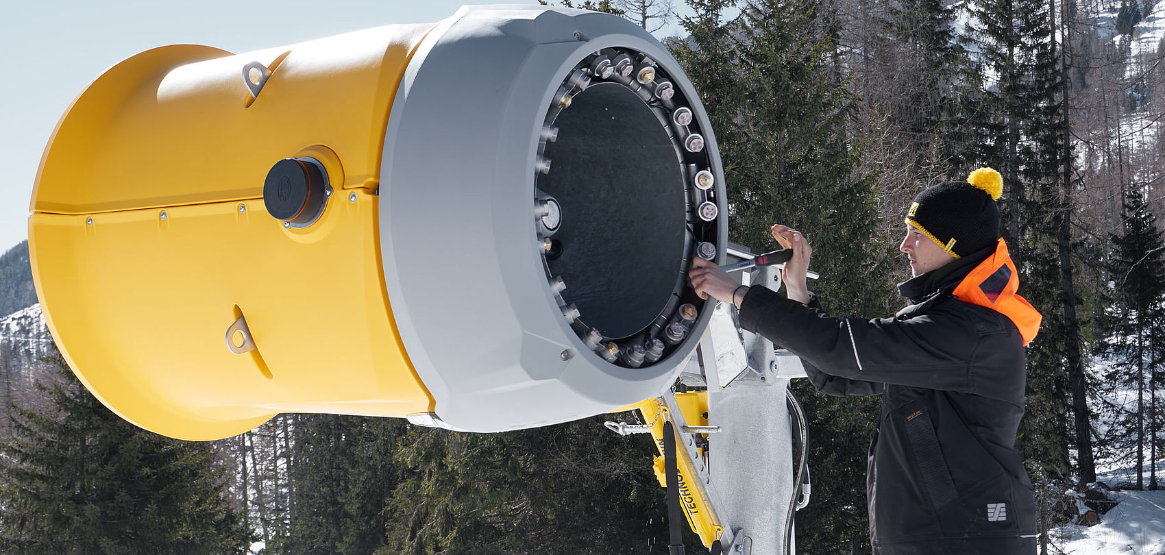 Technician with hat adjusting nozzle ring of yellow TechnoAlpin snow generator outdoors.