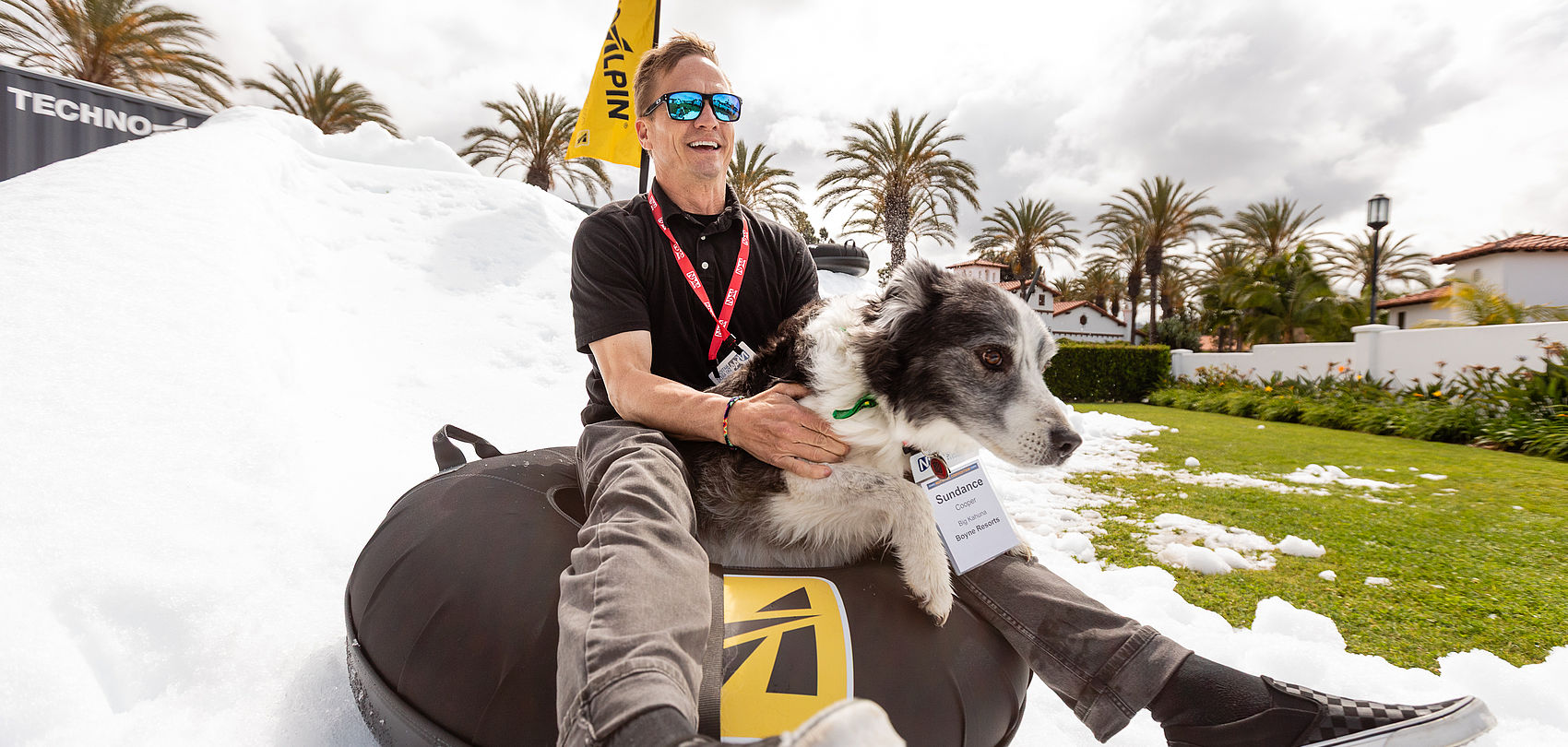 Homme souriant avec lunettes de soleil et chien sur snowtube devant tas de neige et palmiers
