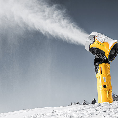 TechnoAlpin TT10 Produktfoto: Hohe gelbe Propellerschneekanone produziert kraftvolle Schneewolke gegen grau-blauen Himmel